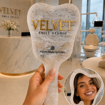 Hand holding a 'Velvet Smile Studio' mirror with a woman's smile inset, in a dental office setting.